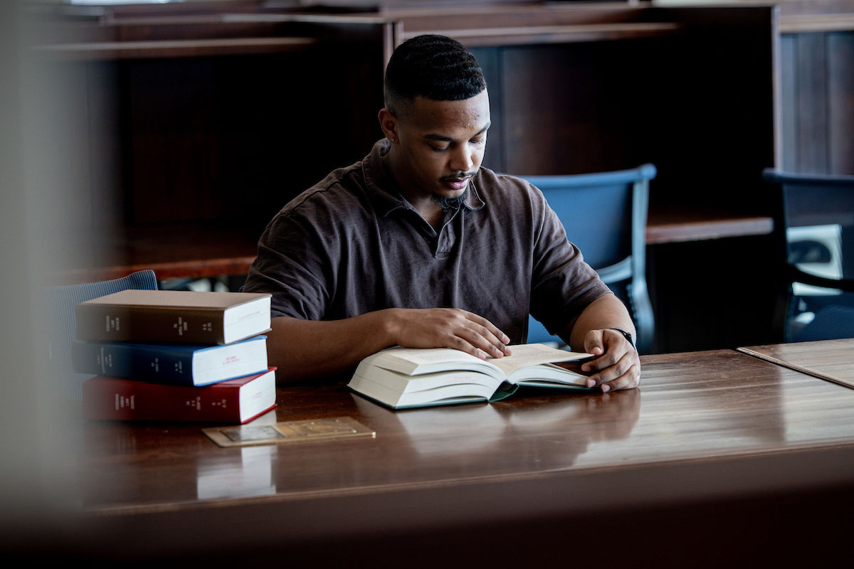 A student sits at a table in the library reading a book with a stack of three books next to him.