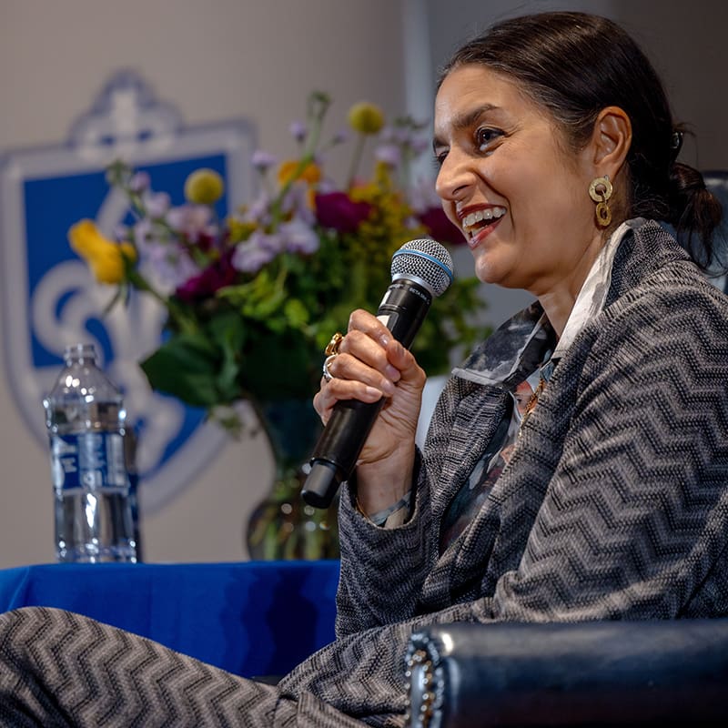 Author Jhumpa Lahiri, wearing a suit, smiles while speaking into a microphone. She sits in a leather chair next to a table with flowers and a water pitcher with the SLU logo in the background.