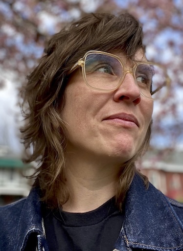 Curator Jessica Baran, wearing a blue jean jacket and large-framed eyeglasses, grins as she looks up and to the right