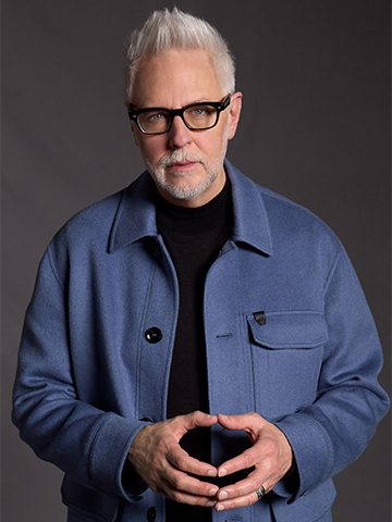 James Gunn, with short, spiked white hair and glasses, wearing a blue jacket over a black shirt, poses thoughtfully with his hands loosely clasped against a dark gray background.    