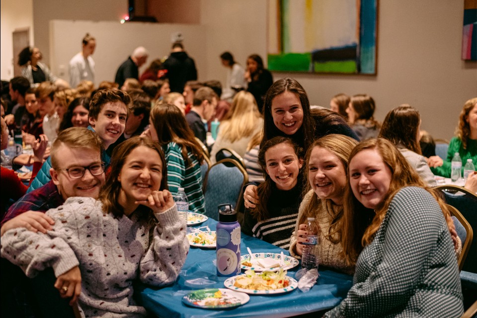 campion society Students smile at the camera while sitting at a long dining table with plates of food.
